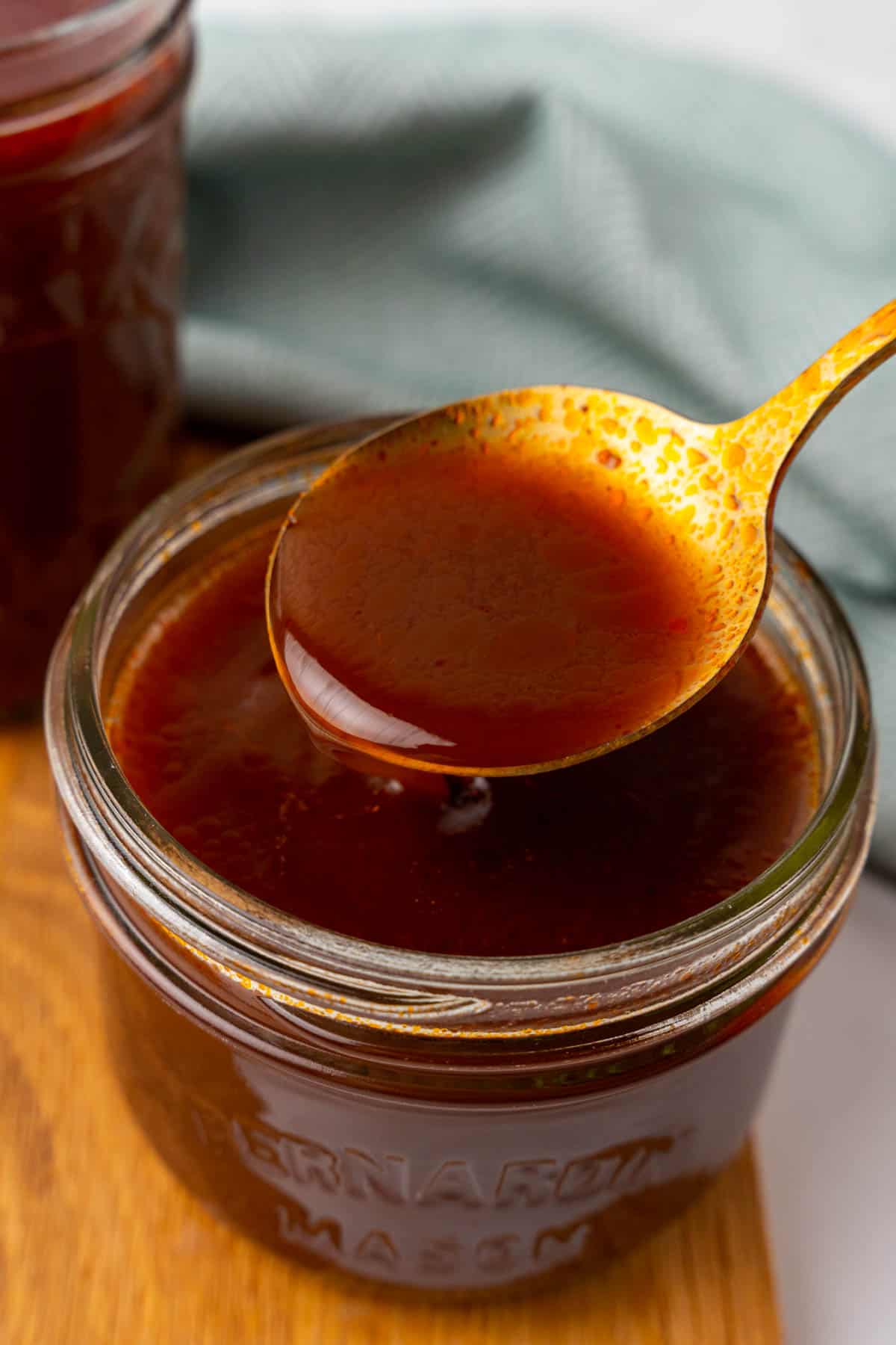 Golden spoon of brown sauce held over short mason jar with more sauce in it on a wooden serving tray