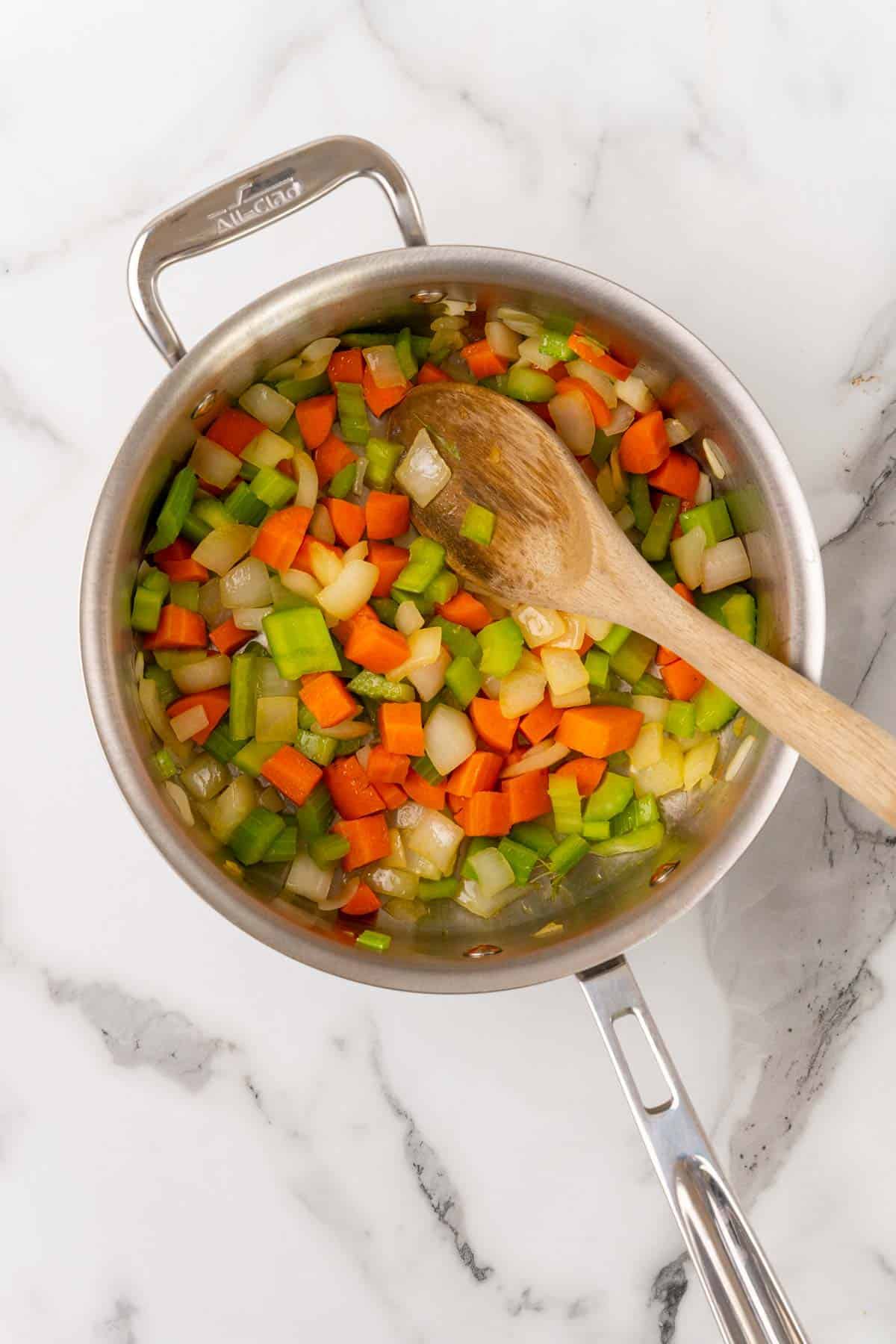 Chopped vegetables added to a silver saucepan with a wooden spoon for stirring, as seen from above on a white marble countertop