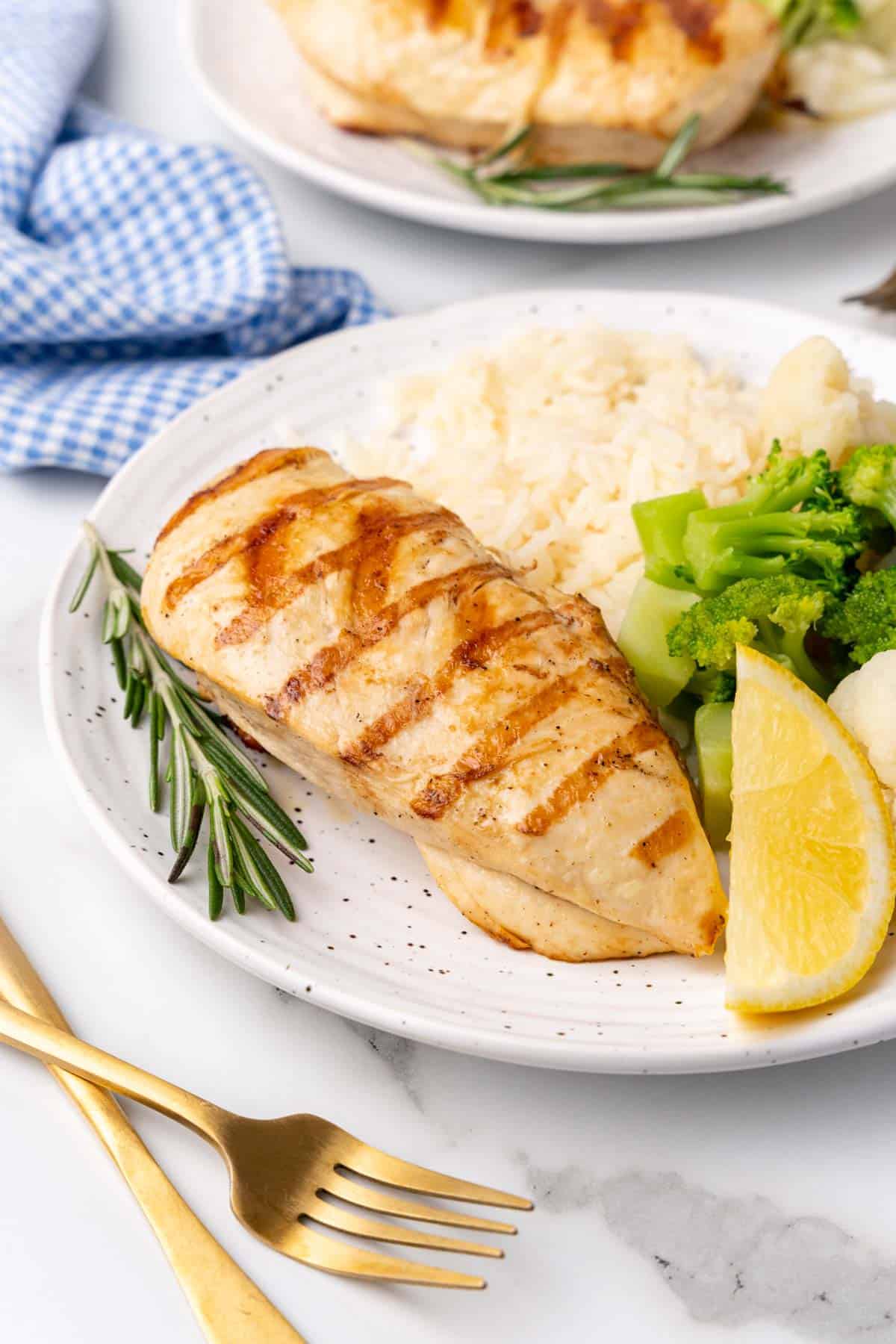 Side angle of chicken breast on a white speckled plate next to a rosemary sprig, lemon wedge, steamed broccoli, and mashed potatoes; golden fork to the side of the plate