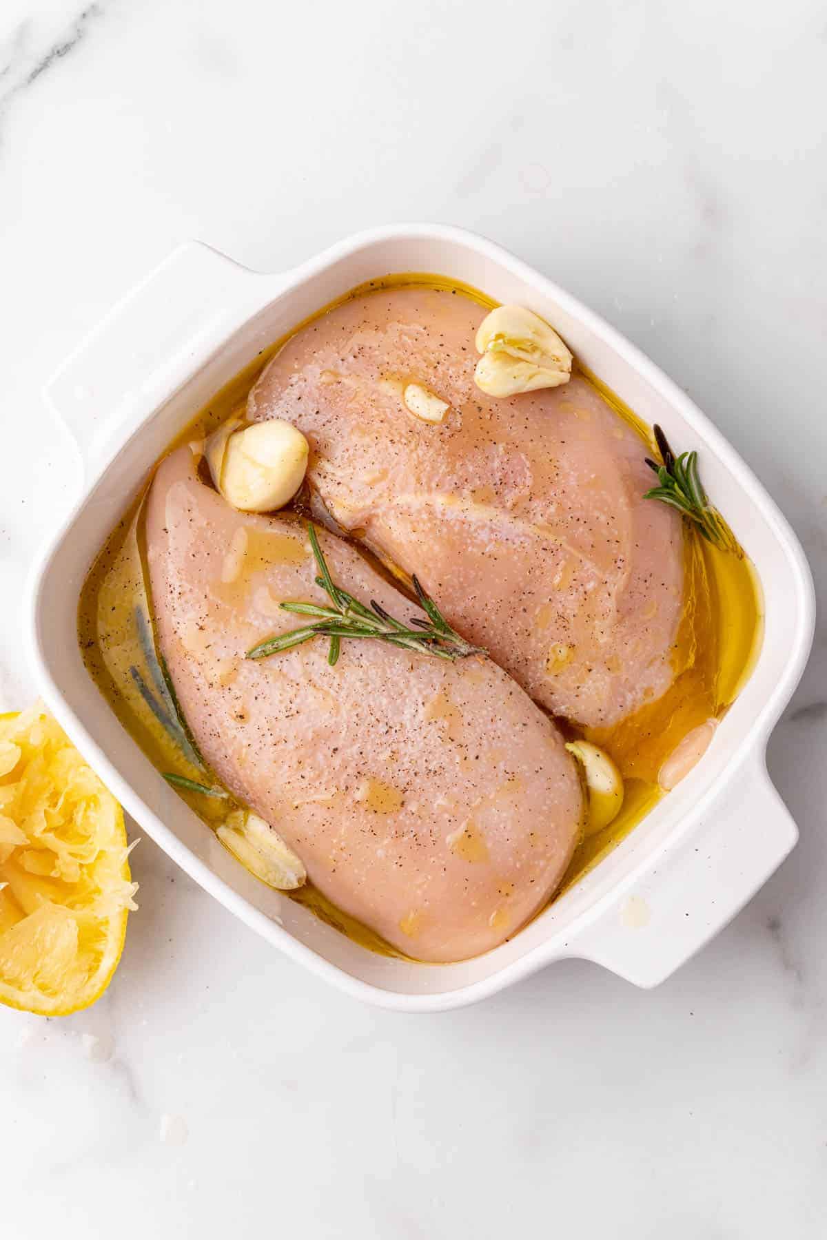 Garlic, rosemary, seasoning, and lemon added to the raw chicken breasts in a white baking dish, as seen from above