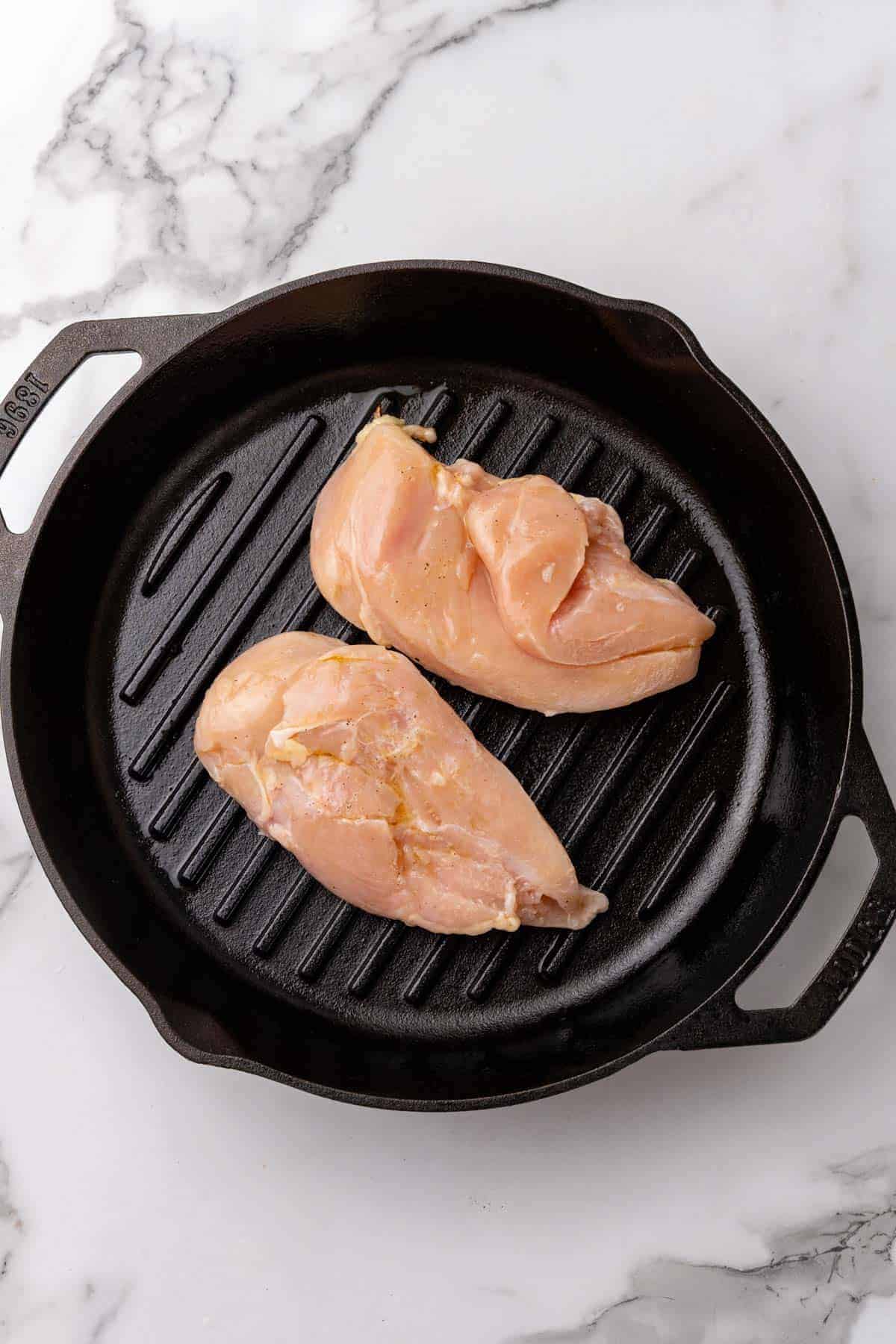 Chicken breasts placed on a black grill pan on a white marble background, as seen from above