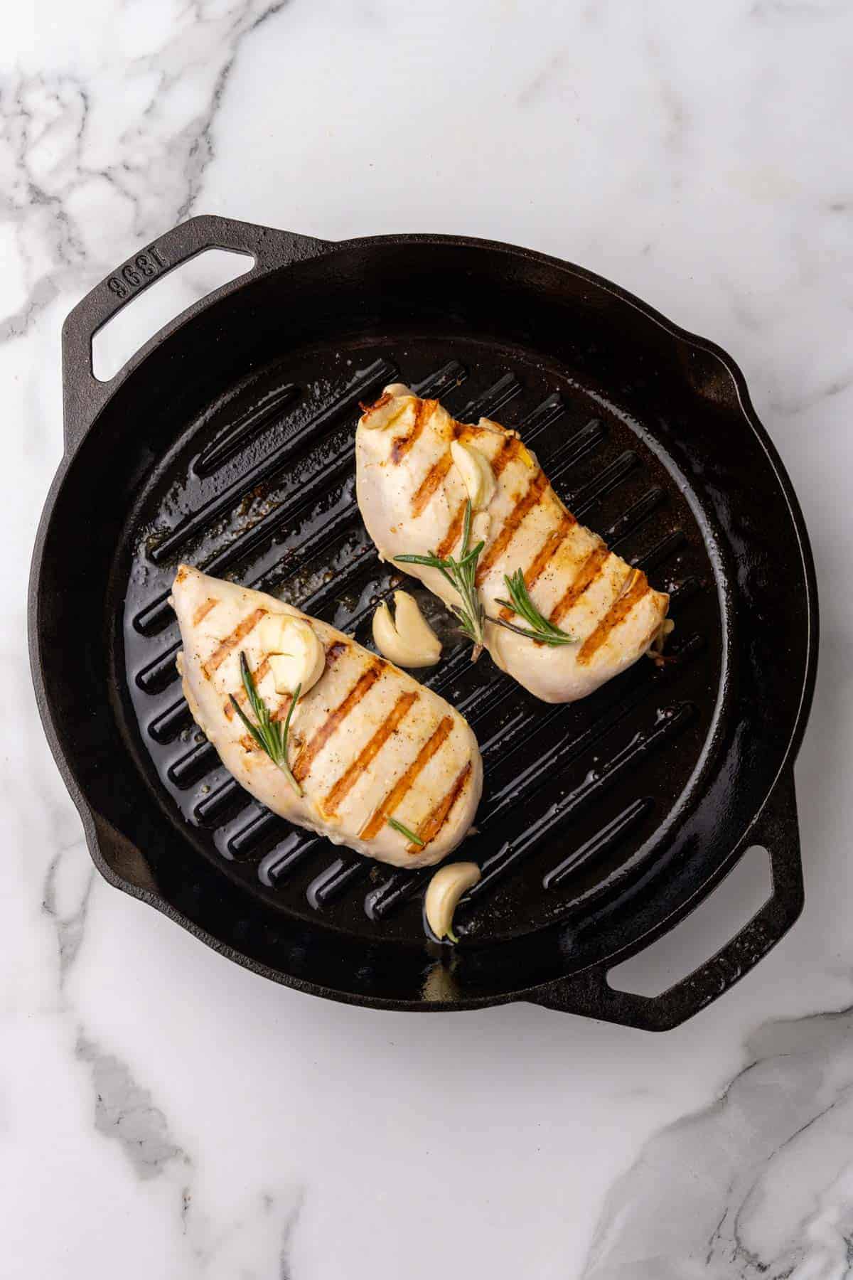 Chicken breasts flipped on the black grill plan so yo can see the grill marks on the side facing up; garlic cloves and rosemary sprigs on top