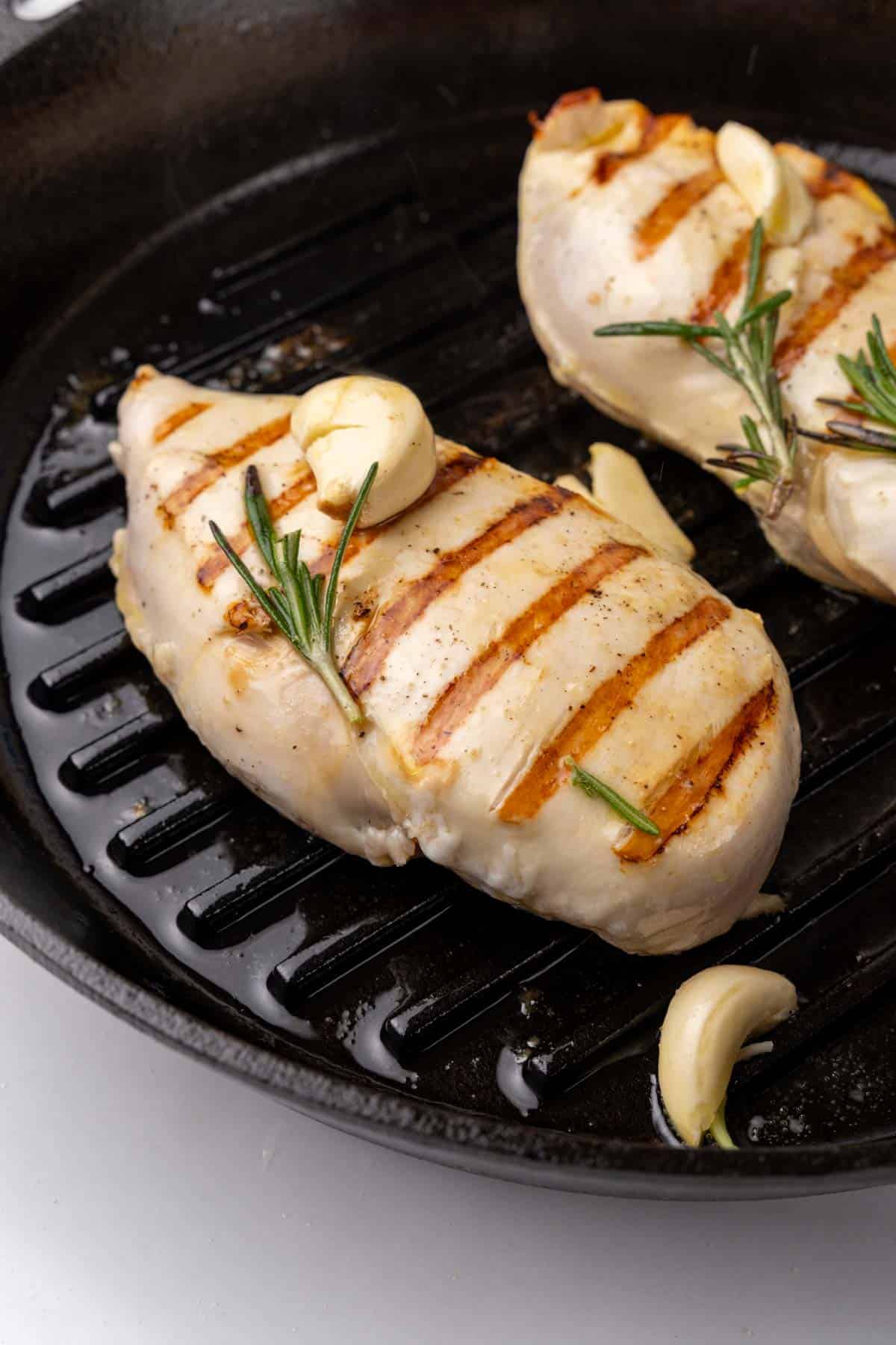 Side view of chicken breasts with grill marks in the black grill pan with rosemary sprigs and garlic cloves on top