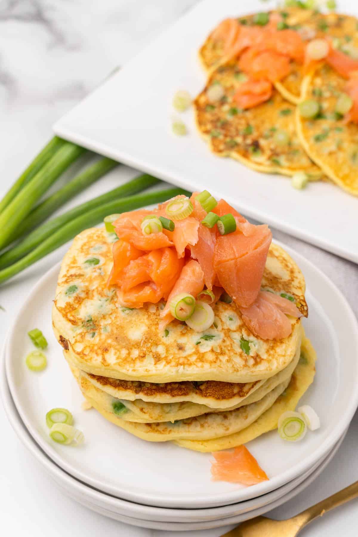 Stack of pea pancakes topped with smoked salmon and sliced scallions on top of a stack of three small white plates with a rectangular white serving platter with more pancakes in the background