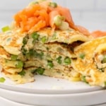 Closeup of a stack of pancakes topped with smoked salmon and sliced scallions with a wedge cut out of the stack