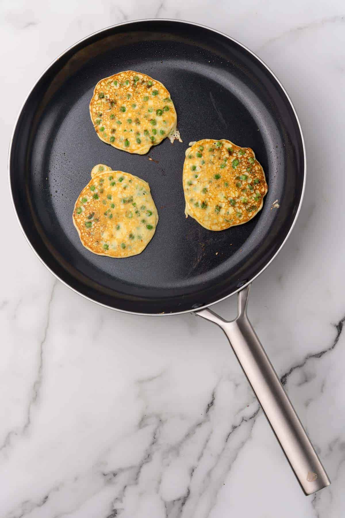 Three pancakes flipped in a black skillet with the golden-brown side facing up, as seen from above on a white marble surface