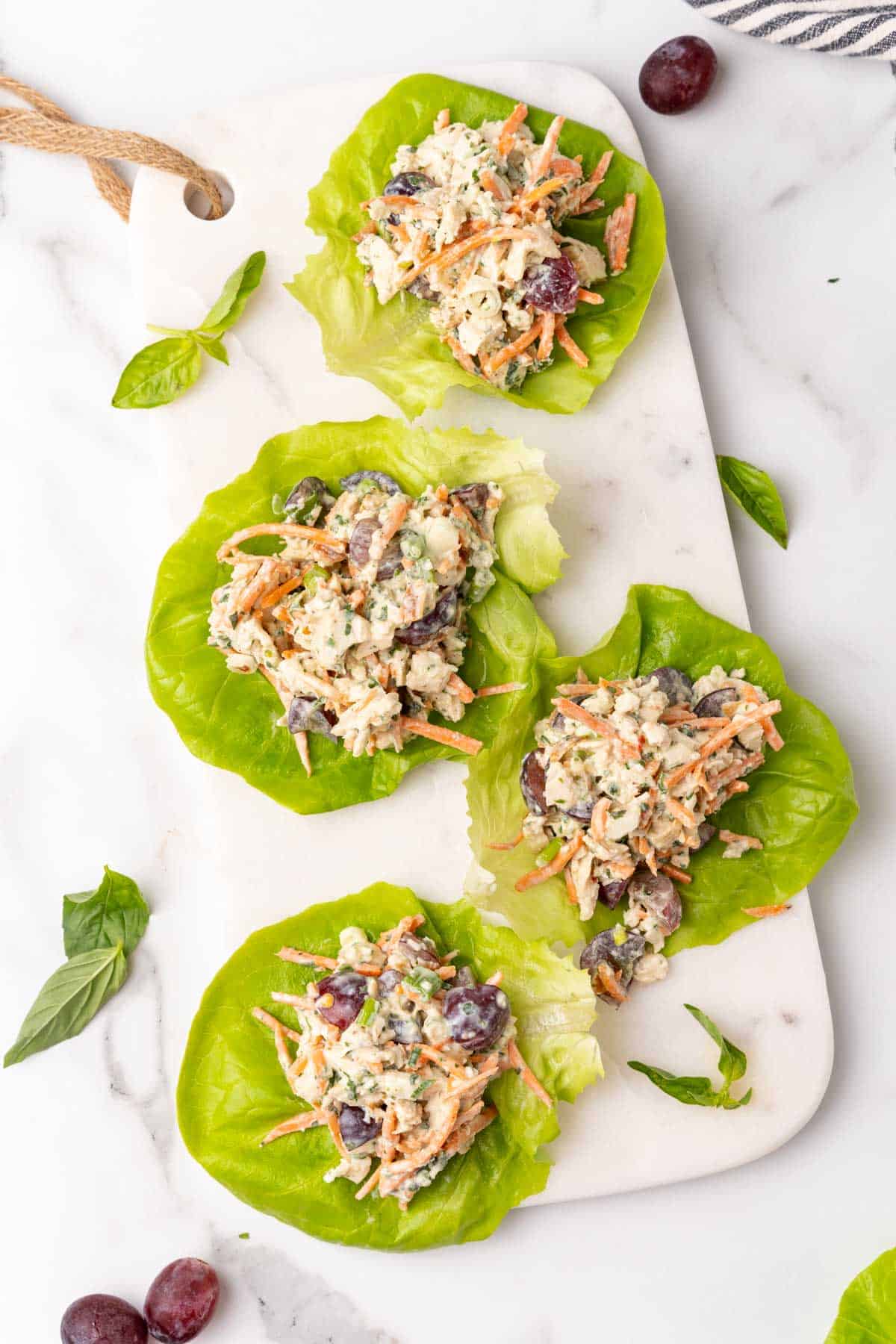 Four lettuce leaves with pesto chicken salad with grapes spooned into the middle of each one, arranged on a white serving tray on a white marble background