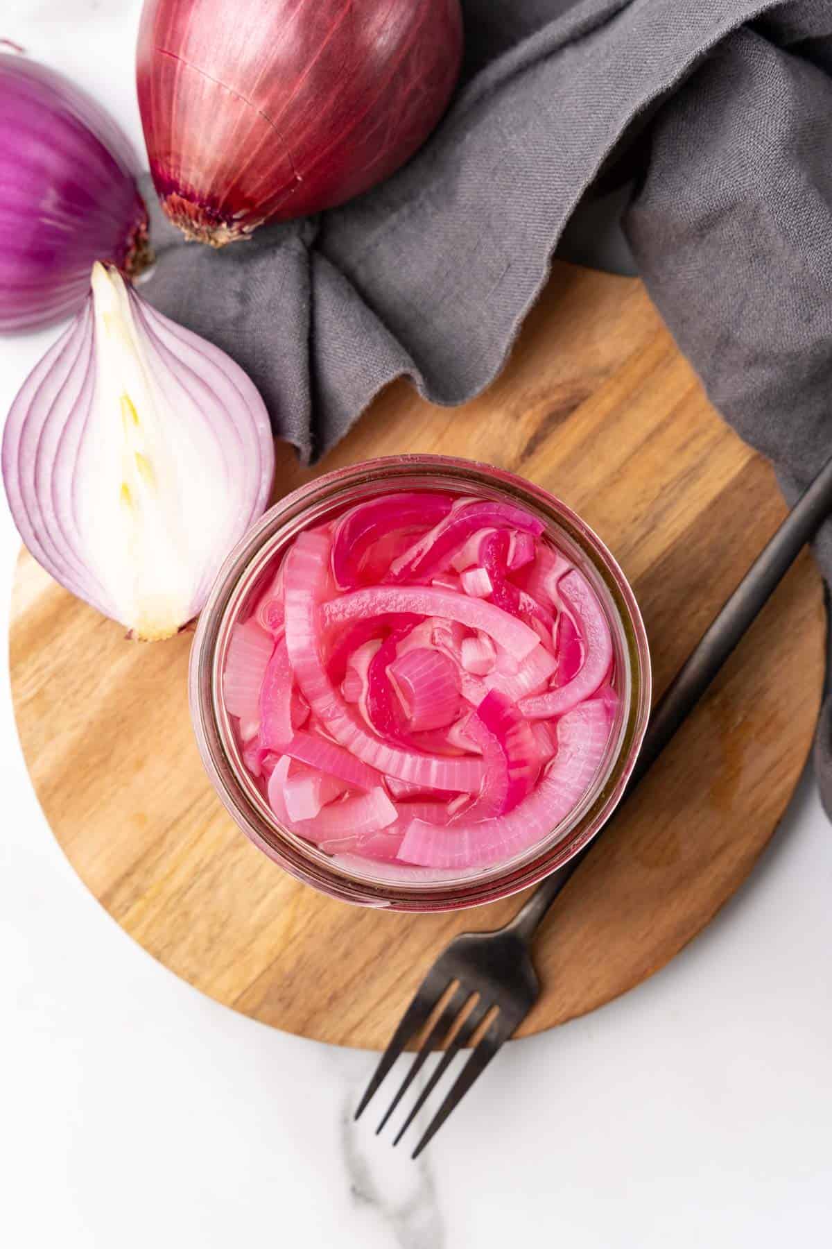 Overhead view of relish in a glass jar on a wooden serving tray next to a metal fork and half of a red onion