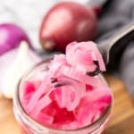 A metal fork lifting pickled pink onion relish out of a glass jar on a wooden serving tray