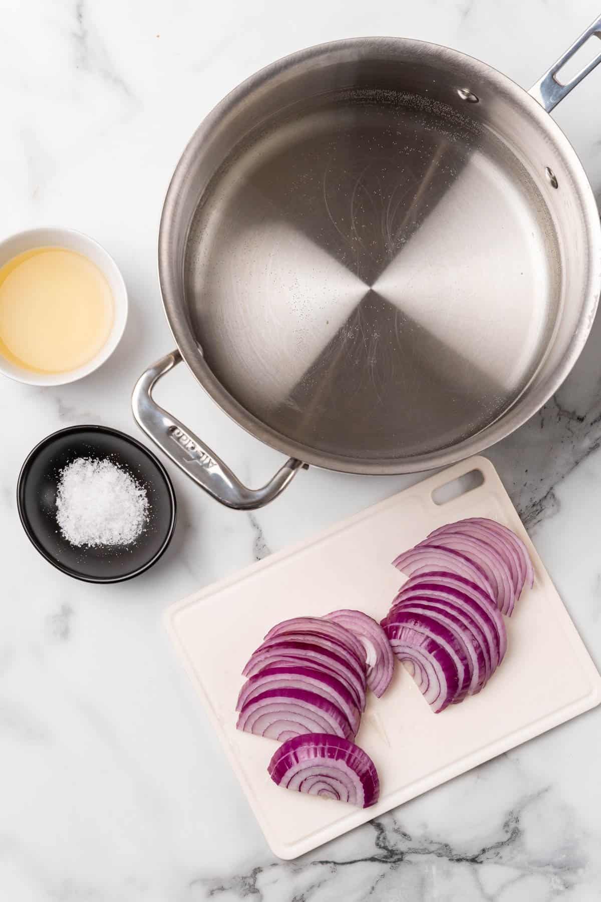 Overhead view of a large metal pot, vinegar in a white ramekin, salt in a black ramekin, and red onion cut into slices on a white cutting board; all seen from above on a white marble surface
