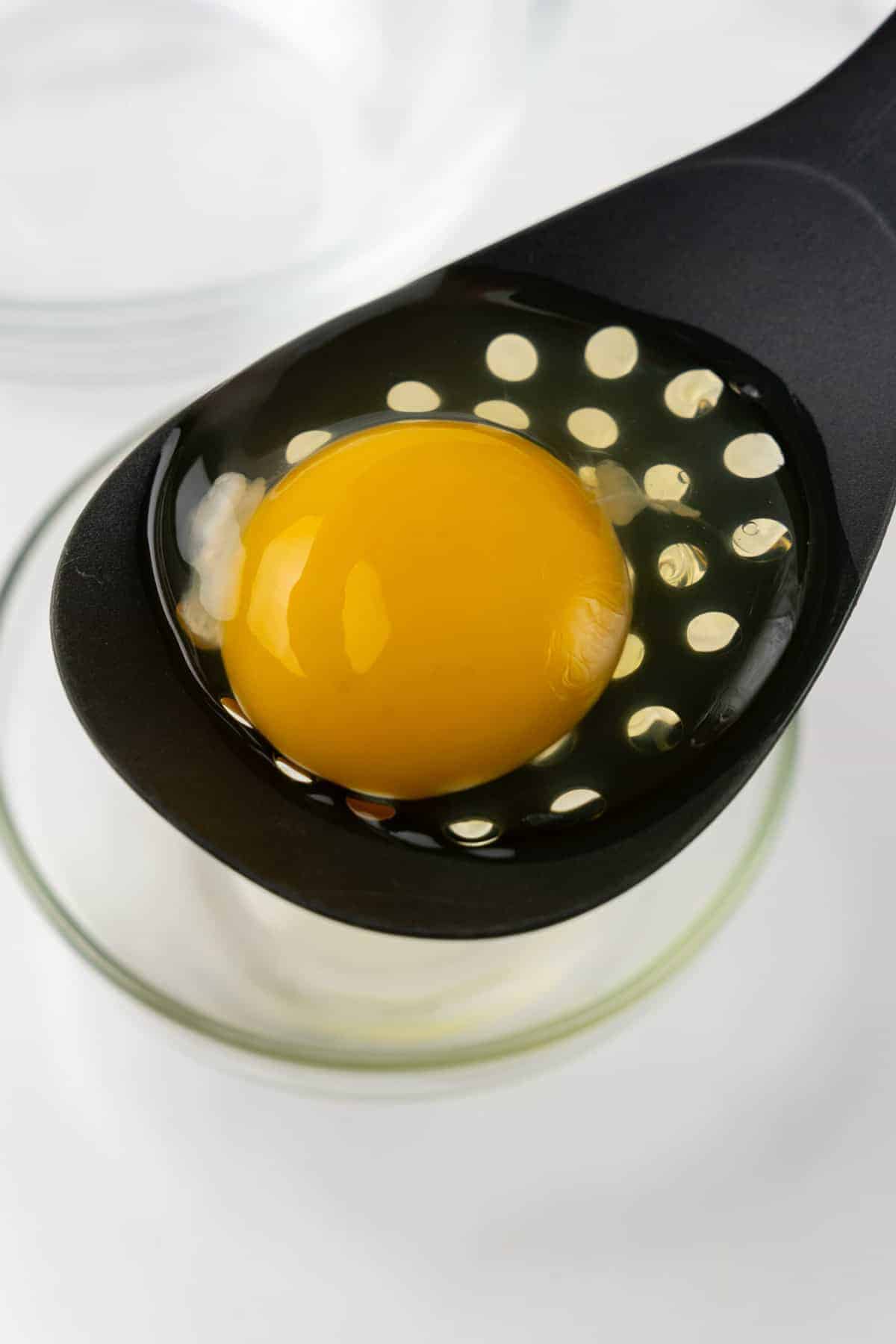 Egg cracked into a black slotted spoon to allow the thin egg whites to drip into a glass ramekin below