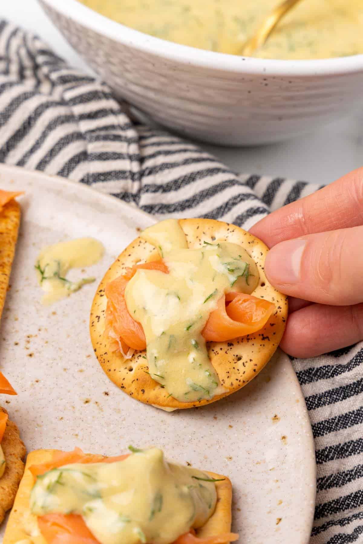 Closeup of hand grabbing a cracker topped with salmon and mustard sauce on a white speckled plate