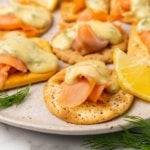 Closeup of crackers topped with salmon and mustard sauce on a white speckled plate with lemon wedges and fresh dill