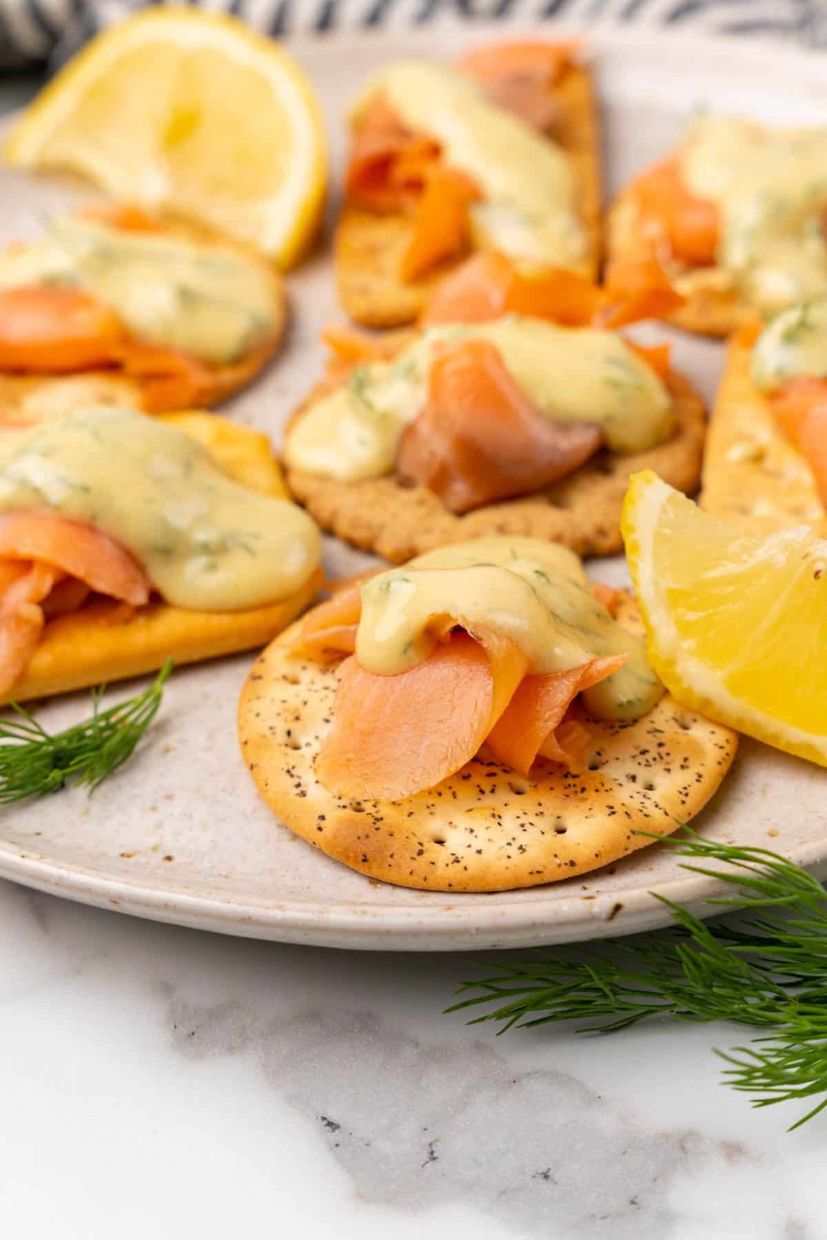 Closeup of crackers topped with salmon and mustard sauce on a white speckled plate with lemon wedges and fresh dill