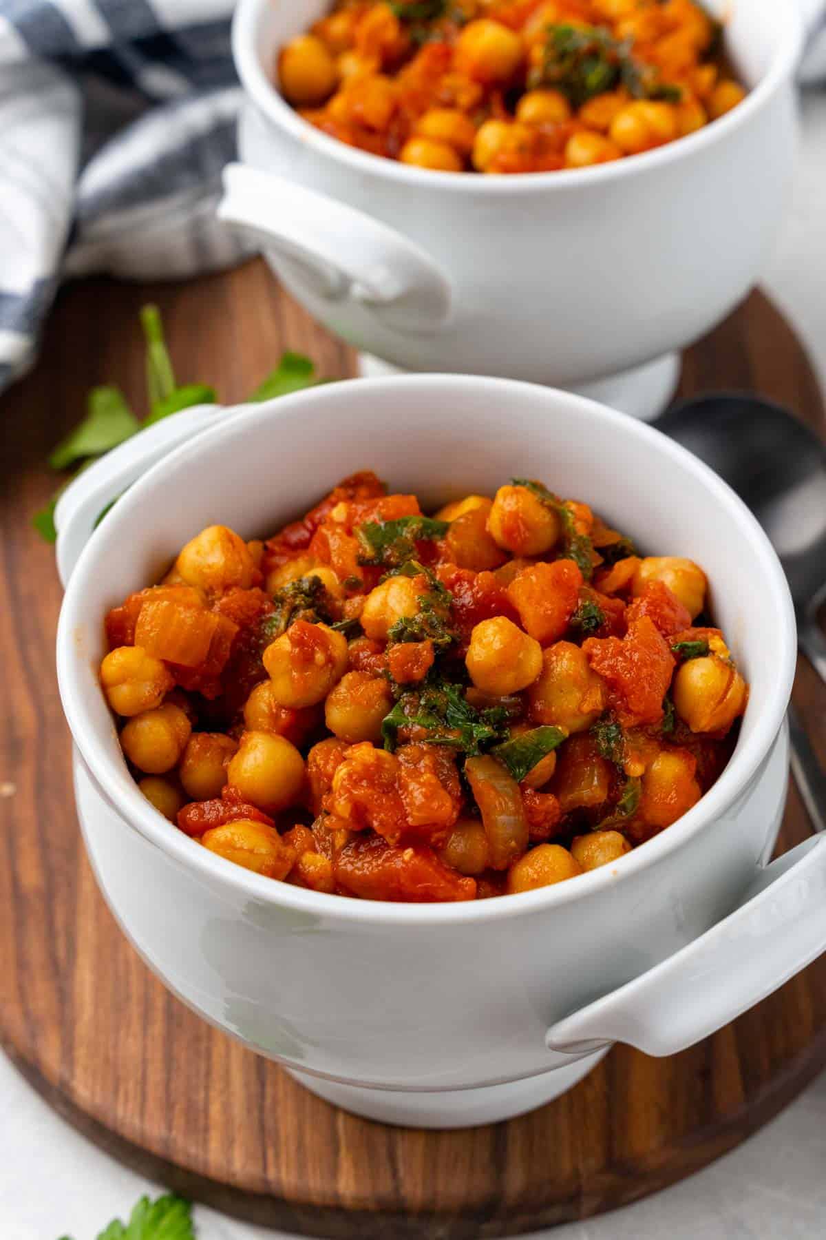 Closeup of stewed chickpeas and tomatoes with kale in a small white bowl with handles on a wooden serving tray with a second bowl and blue striped cloth napkin in the background