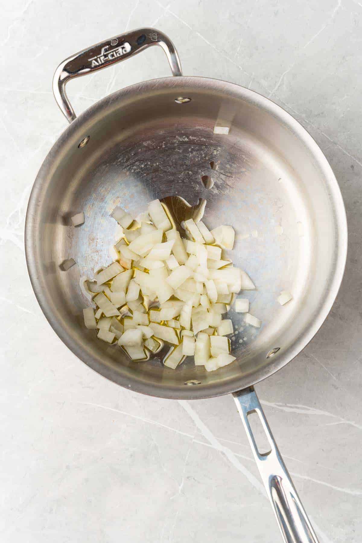 Onions and oil in a silver saucepan on a white marble countertop, as seen form above