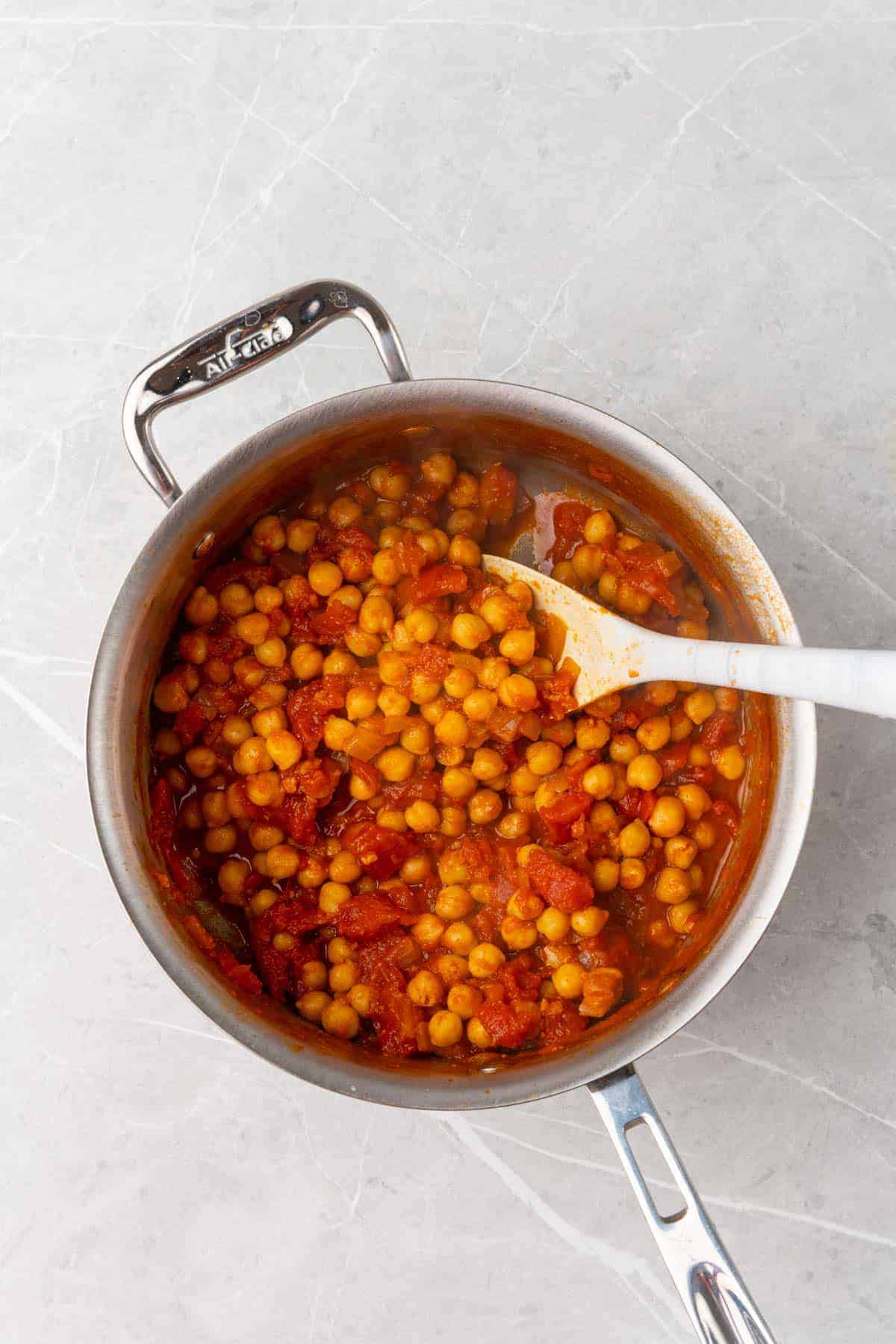 Chickpeas and tomatoes added to the silver saucepan with a white ladle for stirring, as seen from above on a white marble countertop