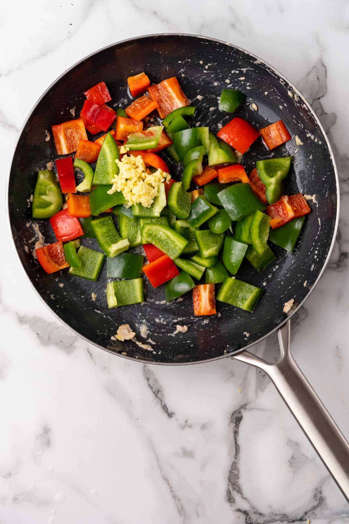 Vegetables and ginger added to black skillet, as seen from above on a white marble surface