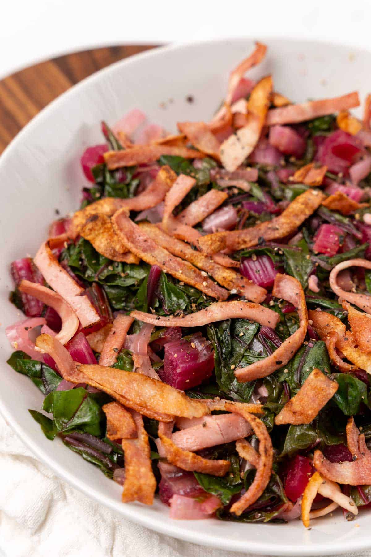Sweet and sour swiss chard topped with turkey bacon in a white bowl on an oval serving tray
