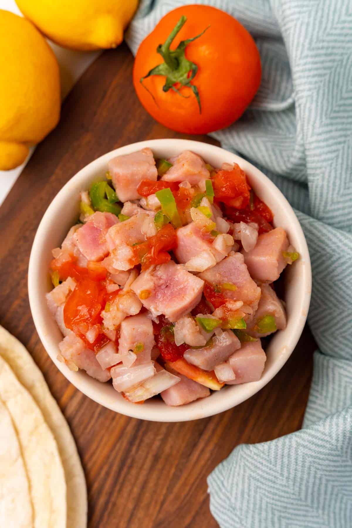 Ceviche in a white bowl as seen from above on a wooden serving board next to a blue cloth napkin and a tomato