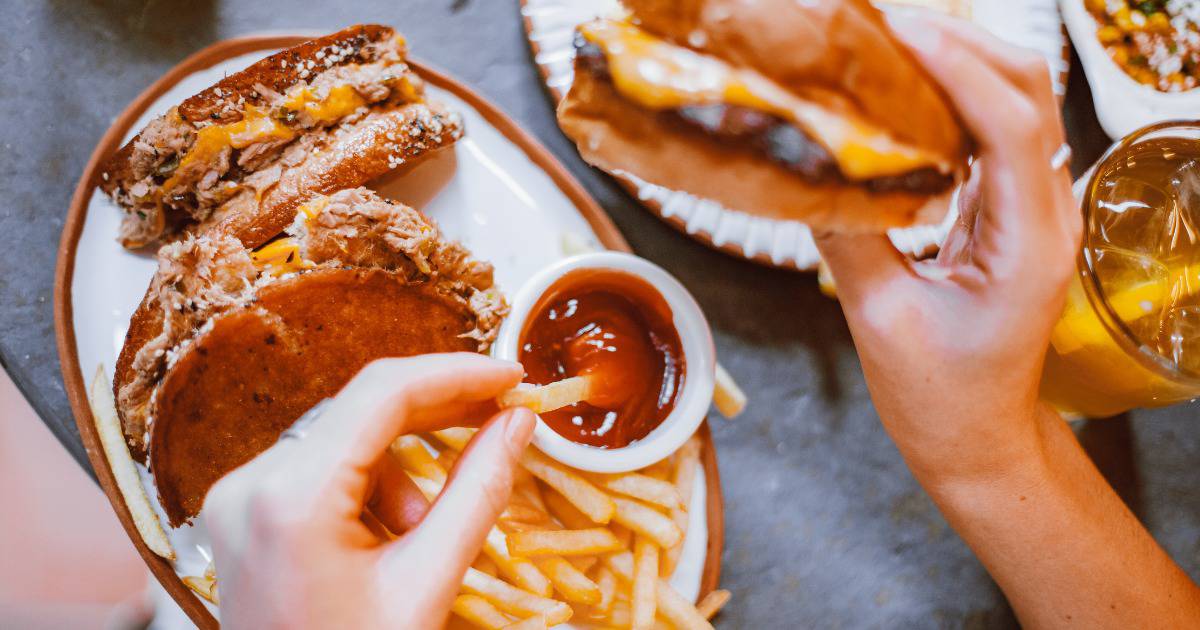 Image of a person dipping a french fry in ketchup with one hand and holding a cheese burger in another hand.