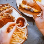 Image of a person dipping a french fry in ketchup with one hand and holding a cheese burger in another hand.