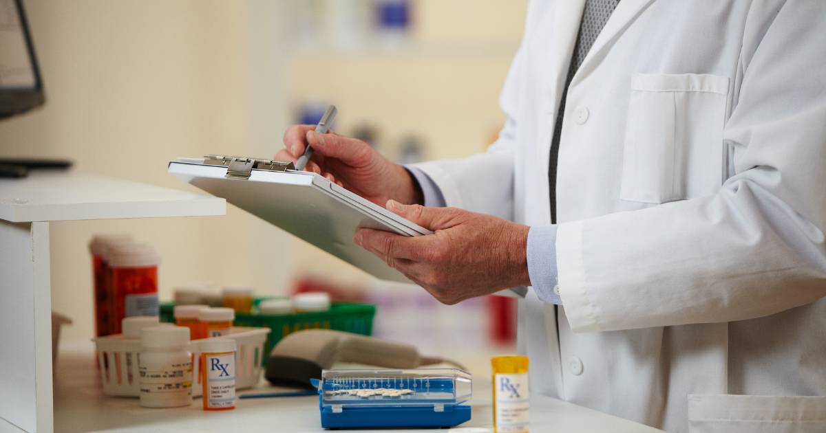 A person in a white lab coat holds a clipboard in front of a counter top with prescription bottles on it.