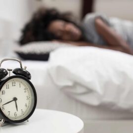 Close-up photo of an alarm clock and a woman sleeping in the background.