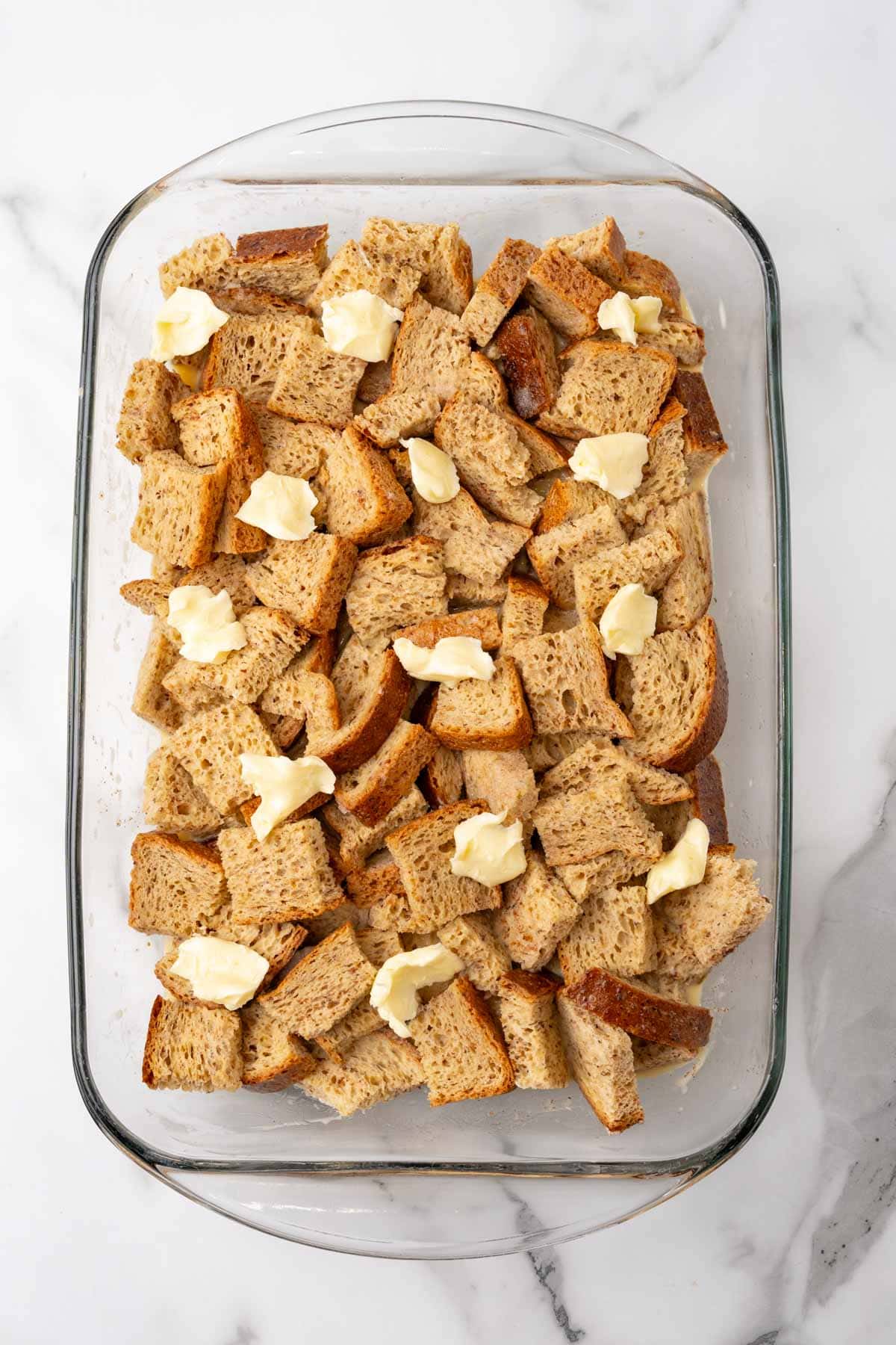 Overhead view of bread slices and butter in a glass baking dish on a white counter